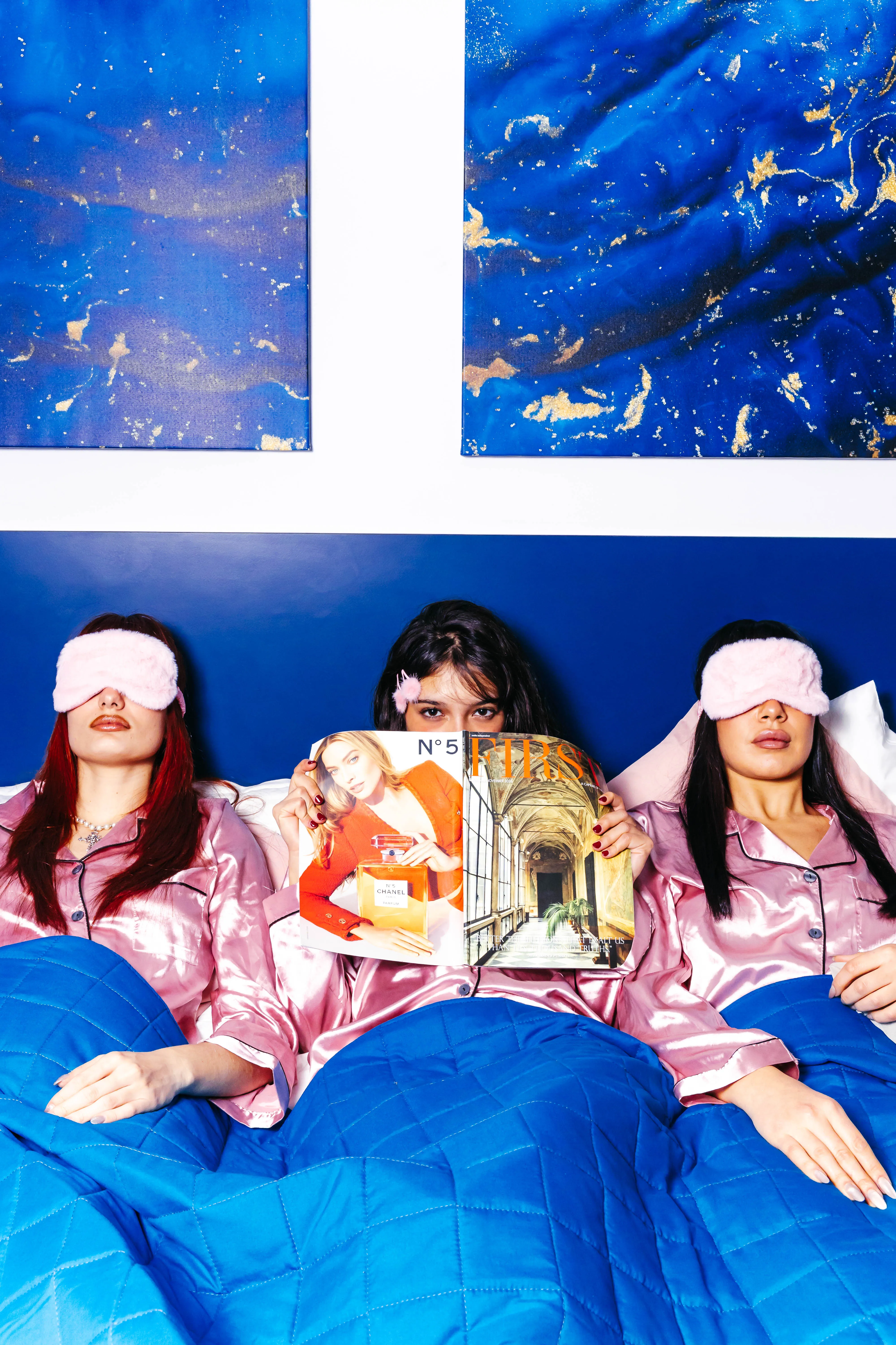 Three friends in pink satin pyjamas reading magazines on a deep blue duvet